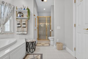 Elegant bathroom with a white tub, glass shower with gold trim, toilet, shelf with towels and plants, window with floral curtains, and sea-themed rugs on a tiled floor. A small stool and wicker trash bin sit nearby.