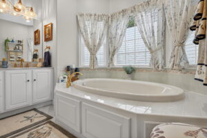 Bright bathroom with a large oval bathtub, white tile surround, gold fixtures, floral curtains on windows, white cabinetry, decorative rugs, and various toiletries and decor on shelves and counter.