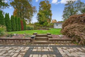 A landscaped backyard with a stone patio, wide steps leading up to a grassy lawn, trimmed shrubs, ornamental grasses, trees, and a white shed in the background under a blue sky.