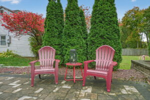 Two red patio chairs sit on a stone patio with a small red table holding a lantern between them. Green shrubs and a tree with red leaves are in the background, along with a grassy yard.