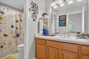 A bright bathroom with a wooden vanity, double sinks, and a large mirror. The shower has a floral and butterfly curtain. Towels are neatly stacked on shelves, and wall art decorates the space.
