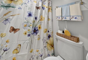 A bathroom with a white toilet beneath a towel rack holding folded towels. The shower curtain features colorful flowers and butterflies. A wicker basket on the toilet tank holds a sponge, tissues, and a toilet paper roll.