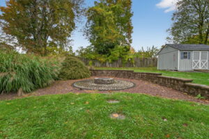 A circular stone fire pit surrounded by a cobblestone patio, bordered by low stone walls, with grass, shrubs, trees, and a gray storage shed in the background under a clear sky.