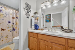 A bathroom with a floral shower curtain, wooden vanity with double sinks, large mirror, wall lamp, white shelves with towels, and decorative items. The walls are light-colored and there are framed pictures above the towel rack.