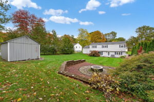 Spacious backyard with green lawn, trees, a circular stone firepit area, a gray shed on the left, and a white two-story house in the background under a blue sky with clouds.