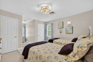 A bedroom with two twin beds covered in yellow floral bedding and purple pillows, light beige walls, framed art above the beds, a large ceiling light, white doors, and windows letting in natural light.