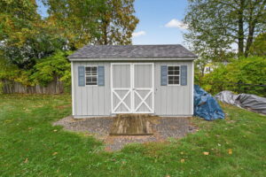A gray backyard shed with double doors, white trim, and blue shutters stands on a gravel patch surrounded by grass and trees on a sunny day. A small wooden ramp leads to the entrance.