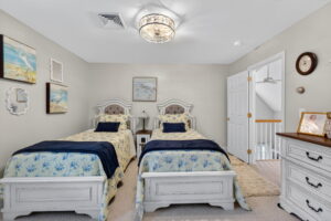 A cozy bedroom with two twin beds featuring blue and yellow floral bedding, matching headboards, a dresser with a clock and framed photo, and beach-themed artwork on soft beige walls under a modern ceiling light.