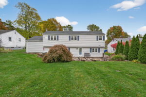 A two-story, light-colored suburban house with dark shutters, a large green lawn, bushes, and trees with autumn foliage under a blue sky with scattered clouds.