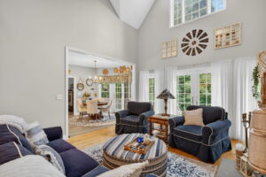 Bright living room with navy and white furniture, round ottoman, and large windows with sheer curtains. Adjacent dining area with a table and chairs, decorated with wall art and clocks. Hardwood floors throughout.