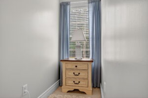 A small cream-colored nightstand with three drawers and a table lamp sits in front of a window with blue curtains. Sunlight shines through the blinds, and the walls are painted white.