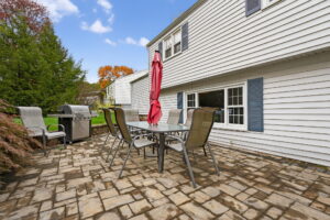 A backyard patio with a glass table, six chairs, a closed red umbrella, and a grill, set on stone pavers outside a white house with blue shutters, surrounded by greenery and autumn trees.