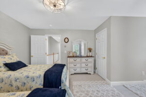 A bedroom with two beds that have yellow and blue floral bedding, a white dresser with a mirror, and neutral-colored walls and carpet. There are two white doors and a framed photo and decor on the dresser.