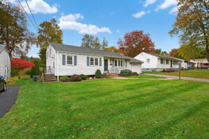A single-story white suburban house with gray shutters, a front porch, driveway, attached garage, and a large green lawn. The yard is bordered by bushes and trees with autumn foliage under a blue sky.