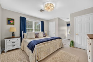 A cozy bedroom with a large bed, white frame, patterned bedding, and blue accents. There are two white nightstands, a white dresser, a potted plant, and windows with blue curtains letting in natural light.