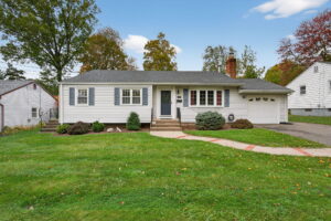 Single-story white house with gray shutters, a dark front door, attached garage, and a well-kept lawn. A concrete path leads to the entrance; there are trees and other houses in the background under a blue sky.
