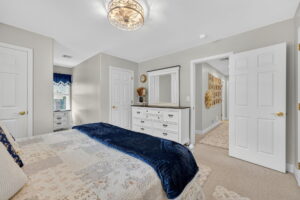 Bright bedroom with a bed featuring a blue throw blanket, a white dresser with a mirror, beige walls, light carpet, and multiple doors leading to a hallway and closets. A decorative ceiling light is overhead.