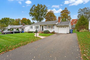 Single-story white house with attached garage, black shutters, and a wide driveway. Green lawn with some fallen leaves, trees with autumn foliage, and neighboring houses visible under a blue sky with clouds.