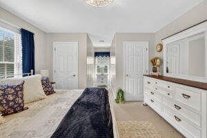 Bright bedroom with a bed on the left, white dresser with mirror on the right, two doors, beige walls, blue curtains, and a window at the far end letting in natural light.