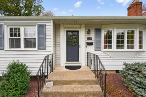 Front entrance of a white house with blue shutters, a gray door decorated with a wreath, black metal railings on both sides of the steps, and plants and shrubs along the walkway. House number 29 is displayed by the door.