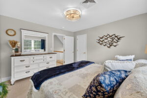 A cozy bedroom with a neatly made bed, patterned pillows, a white dresser with a mirror, wall art of flying birds, and neutral walls; natural light streams in through a window reflected in the mirror.