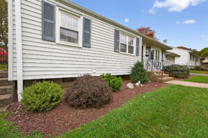 A single-story house with white siding, blue shutters, and a small front garden with bushes and mulch. Concrete steps and a metal handrail lead up to the front door. The sky is blue with a few clouds.