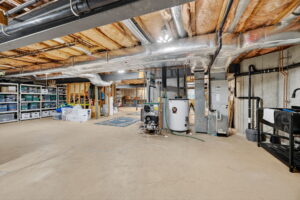 Spacious unfinished basement with exposed ceiling beams, HVAC ducts, a water heater, utility sink, storage shelves, plastic bins, and various household items organized along the walls. Concrete floors and partially finished walls.