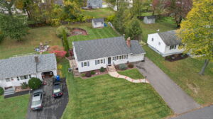 Aerial view of a single-story house with a gray roof, driveway, front lawn, and large backyard with a circular fire pit and two small sheds. Neighboring houses and autumn trees are visible.