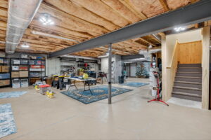 Spacious unfinished basement with exposed ceiling beams, storage shelves filled with boxes, workbenches, tools, two blue rugs, and a staircase leading upstairs. The floor is concrete with visible cracks.
