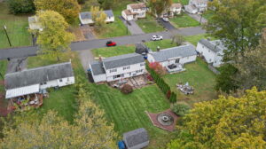 Aerial view of suburban houses with green lawns, trees with autumn foliage, and a small shed and circular garden feature in one backyard. Cars are parked along the residential street.