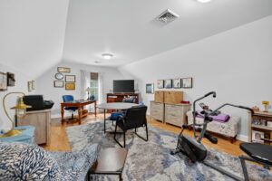 A spacious, well-lit home office and exercise room with a desk, computer, exercise bike, TV, storage units, armchairs, and a patterned rug on a wooden floor. Artwork and decor are displayed on the walls.
