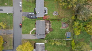 Aerial view of a suburban neighborhood showing houses with gray roofs, green lawns, trees, a driveway with parked cars, a patio area, and a circular garden feature in the backyard.