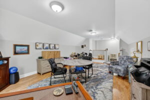 A spacious home office with desks, a round table, chairs, art on the wall, a patterned rug, and wood flooring. The room has sloped ceilings, a cozy sitting area, and modern lighting.