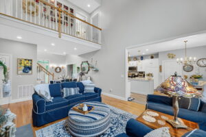 A cozy living room with navy blue sofas, a striped ottoman, and a Tiffany-style lamp. The space features hardwood floors, light gray walls, and an open view into a white kitchen and staircase with a balcony above.