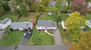 Aerial view of suburban homes with green lawns, driveways, cars parked, and a backyard garden with trees and a small shed. The trees are in autumn foliage with some leaves turning yellow and red.