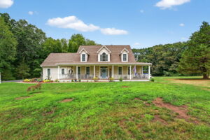 A white, two-story house with dormer windows and a large front porch sits in the middle of a spacious, green lawn bordered by trees under a blue sky with a few clouds.