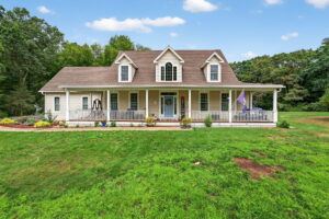 A beige two-story house with white trim, dormer windows, and a large front porch sits on a green lawn, surrounded by trees and colorful flower beds under a blue sky with scattered clouds.