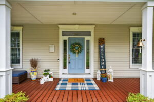 A front porch with a light blue door, a wreath, a striped doormat, and a WELCOME sign. The porch has wooden flooring, white columns, potted plants, lanterns, and decorative items.