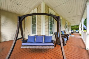 A spacious porch with tan siding features a wooden swing with blue cushions and pillows, wicker chairs with blue cushions, and decorative plants, overlooking a green yard.