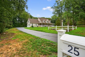 A white house with a wraparound porch sits behind a curved driveway and a white fence. The mailbox in the foreground displays the number 25. The property is surrounded by trees and green grass under a blue sky.