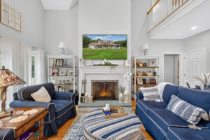 A cozy living room with blue sofas, a striped ottoman, a lit fireplace, and bookshelves on each side. Above the fireplace is a TV displaying a house. The room has high ceilings and a staircase with a balcony.