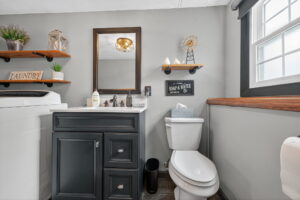 A small bathroom with a gray vanity, white sink, mirror, and toilet. Wooden shelves hold decor and plants. A window lets in natural light. A Soap & Water sign and laundry bin are also visible.