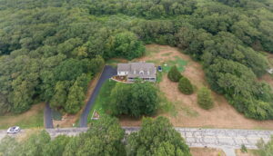 Aerial view of a large house surrounded by trees and a spacious yard, with a winding driveway and a road in front. The property is bordered by dense forest on three sides.