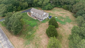 Aerial view of a two-story house with a porch, surrounded by trees and a large grassy yard. A driveway curves to the side of the house, and a single vehicle is parked nearby. Forested area borders the property.