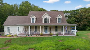 A two-story beige house with a large front porch, blue front door, dormer windows, and a well-maintained green lawn, surrounded by trees under a partly cloudy sky.