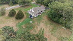 Aerial view of a house with a porch surrounded by trees and a large grassy yard, with a boat parked next to the house and a paved driveway nearby.
