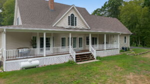 A beige, two-story house with a large covered front porch, white railings, and steps leading to a grassy yard. There is a propane tank on the left and trees in the background.