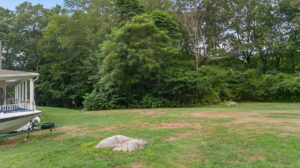 A grassy yard with a large rock in the foreground, bordered by dense green trees. The edge of a house and a boat on a trailer are visible on the left side of the image.