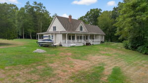 A beige house with a large covered porch sits on a grassy lawn surrounded by trees. A small boat on a trailer is parked next to the house. The sky is partly cloudy.