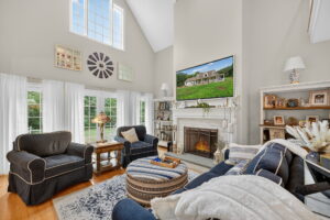 Bright, spacious living room with high ceilings, large windows, and a fireplace. Dark blue sofas, a patterned ottoman, bookshelves, and decor create a cozy, inviting atmosphere. A TV is mounted above the fireplace.